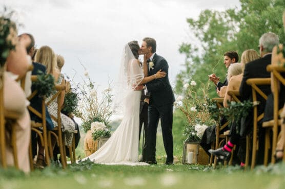 A bride and groom share their first kiss at an outdoor wedding ceremony, surrounded by seated guests on either side, with greenery and floral decorations in the background. by Justin Salem Meyer
