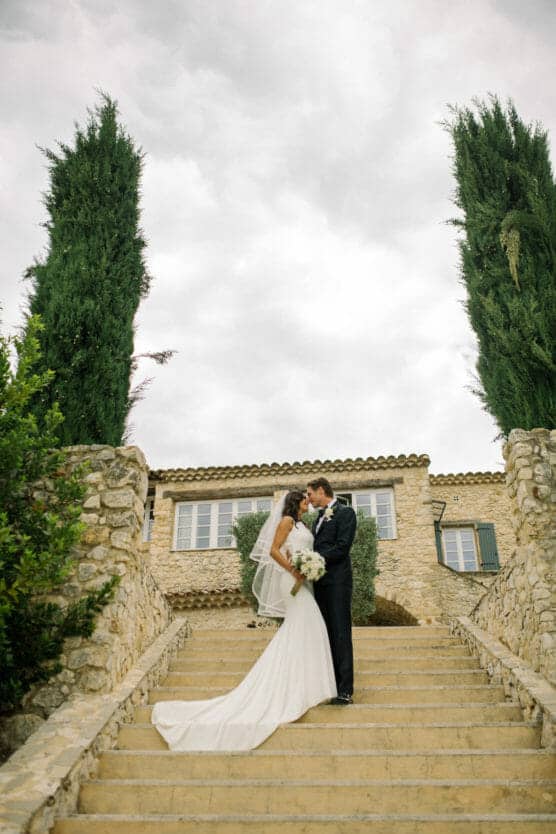 A bride and groom stand on stone steps, embracing and smiling at each other in front of a rustic stone building with green shutters, framed by tall cypress trees under a cloudy sky. by Justin Salem Meyer