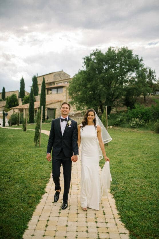 A bride in a white dress and a groom in a black tuxedo walk hand in hand on a stone path through a grassy garden, with rustic buildings and trees in the background under a cloudy sky. by Justin Salem Meyer