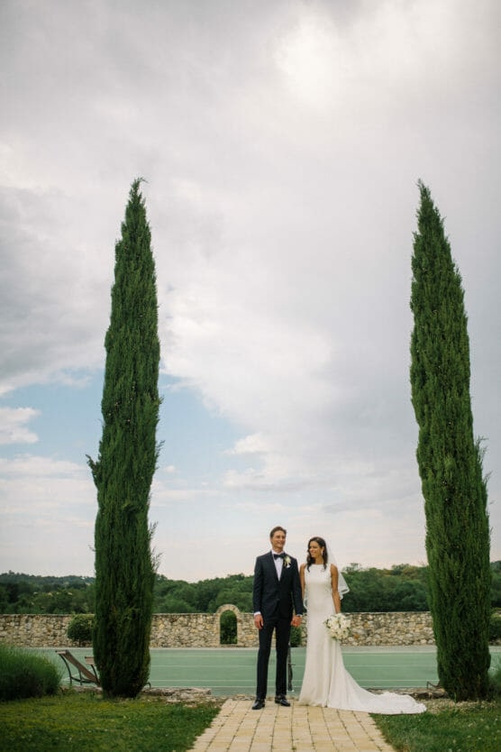 A bride and groom stand holding hands outdoors between two tall cypress trees on a stone pathway, with greenery and a cloudy sky in the background. The bride holds a bouquet and wears a white dress; the groom wears a black suit. by Justin Salem Meyer