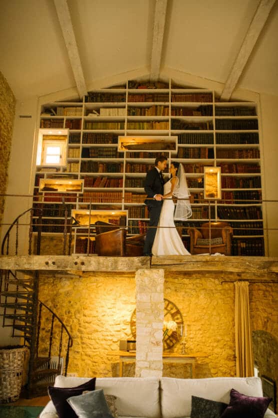A bride and groom stand together on a wooden loft in front of a tall bookshelf filled with books in a cozy, rustic room with stone walls and warm lighting. by Justin Salem Meyer
