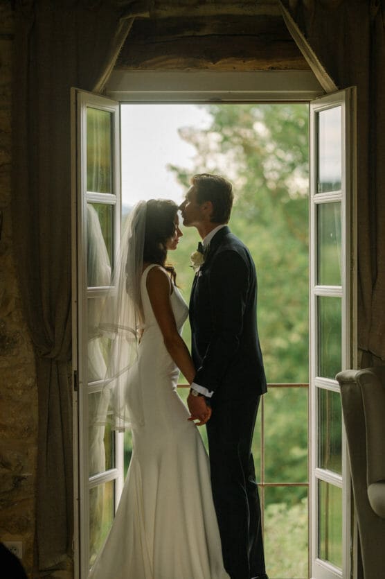 A bride and groom stand in a doorway framed by open French doors. The groom gently kisses the bride’s forehead as they hold hands, with greenery visible outside. Soft natural light filters into the room. by Justin Salem Meyer