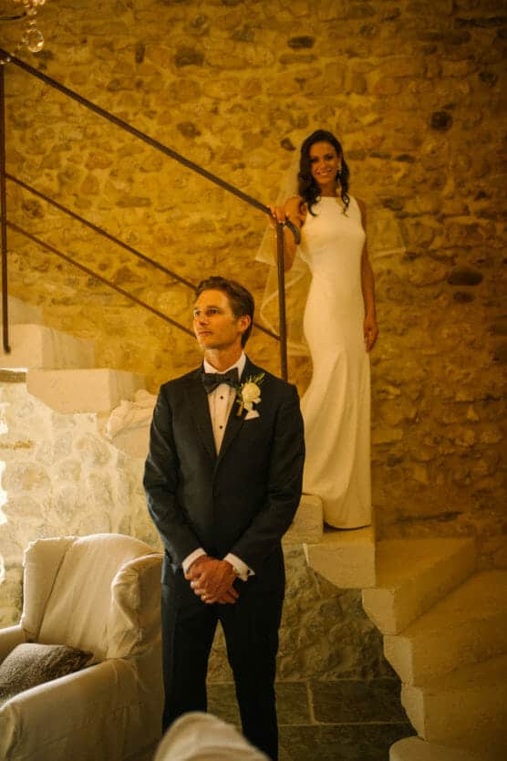 A bride in a white dress stands on stairs behind a groom in a black tuxedo, indoors with stone walls and soft lighting. The groom faces forward while the bride looks at him, both preparing for a special moment. by Justin Salem Meyer
