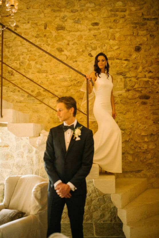 A bride in a white dress stands on a stone staircase, looking at a groom in a black tuxedo who stands in the foreground with his hands folded, both inside a rustic stone-walled room. by Justin Salem Meyer