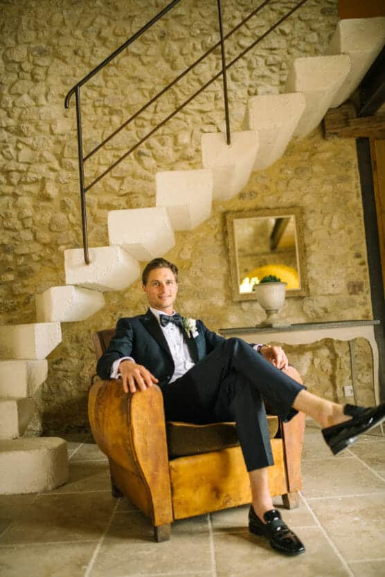 A man in a tuxedo sits confidently in a brown leather armchair, legs crossed, under a white stone staircase in a rustic room with stone walls and tiled floor. A mirror and a potted plant sit on a table in the background. by Justin Salem Meyer