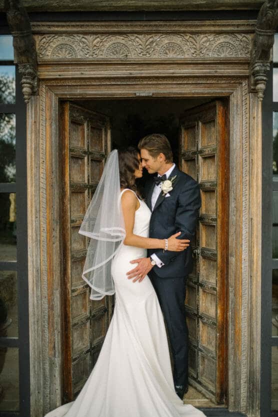 A bride in a white dress and veil embraces and kisses a groom in a dark suit and bow tie as they stand together in a carved wooden doorway. by Justin Salem Meyer