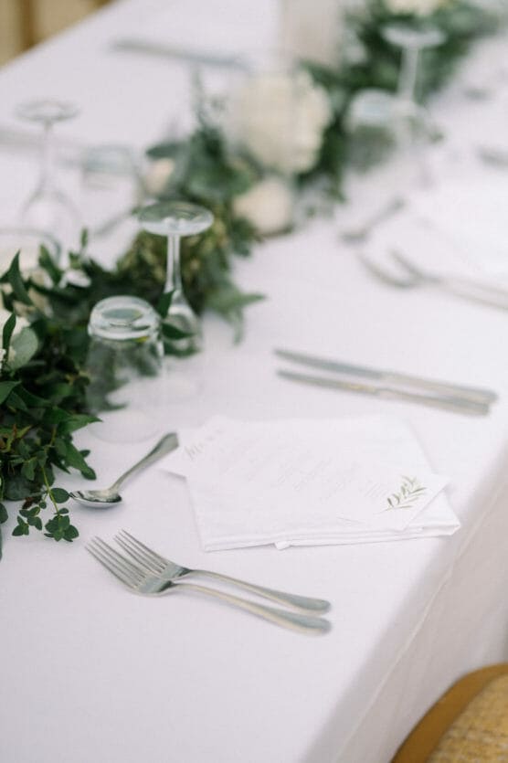 A close-up of a white tablecloth with a formal place setting, including silverware, wine glasses turned upside down, a menu, and green foliage decor running along the center of the table. by Justin Salem Meyer