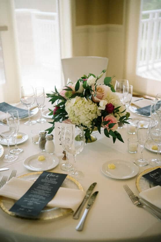 Elegant round table set for a formal event with gold-rimmed plates, neatly folded napkins, menus, glassware, and a centerpiece of white, pink, and burgundy flowers with greenery. Natural light streams in from windows. by Justin Salem Meyer