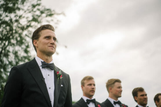 A man in a tuxedo with a bow tie and boutonniere stands outdoors, smiling slightly, with four other men in tuxedos blurred in the background, under a cloudy sky. by Justin Salem Meyer
