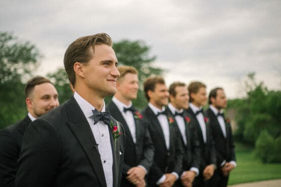 A group of men in black tuxedos with bow ties stand in a row outdoors, smiling and looking forward. Trees and greenery are in the background under a cloudy sky. by Justin Salem Meyer