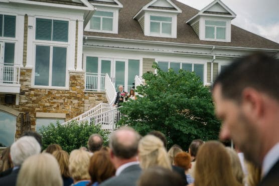 A bride and a man, possibly her father, stand at the top of an outdoor staircase while guests watch from below, in front of a large house with stone and white siding. by Justin Salem Meyer