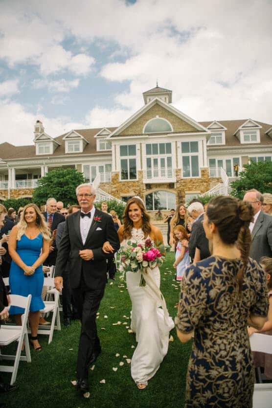 A smiling bride in a white dress holding a bouquet walks down the outdoor aisle with an older man at her side, surrounded by seated and standing guests, in front of a large elegant house. by Justin Salem Meyer