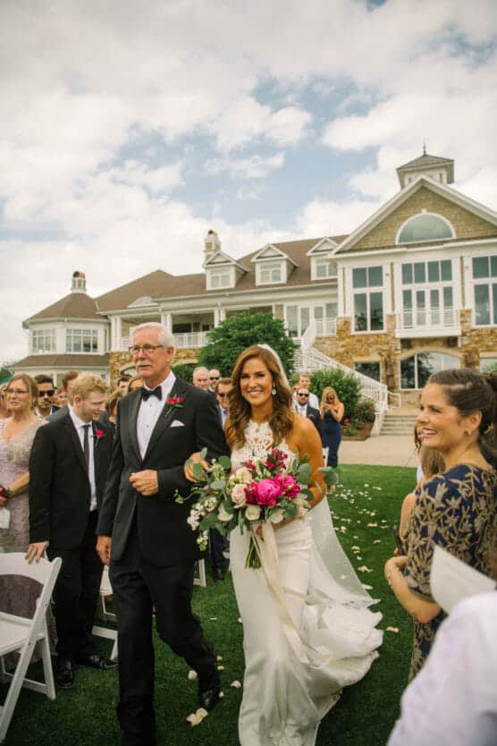 A bride in a white dress and veil holding a vibrant bouquet walks down the outdoor aisle with an older man in a tuxedo, surrounded by smiling guests, in front of a large elegant house under a partly cloudy sky. by Justin Salem Meyer