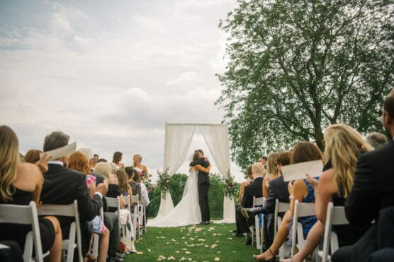 A bride and groom embrace under a white arch during an outdoor wedding ceremony, surrounded by seated guests on either side. Flower petals are scattered on the grass aisle, and a large tree stands nearby under a partly cloudy sky. by Justin Salem Meyer