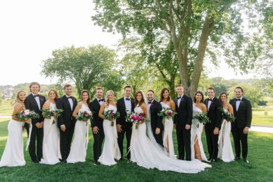 A wedding party poses outdoors on grass with trees in the background. The bridesmaids wear white dresses and hold bouquets, while the groomsmen wear black suits with bow ties. Everyone is smiling at the camera. by Justin Salem Meyer