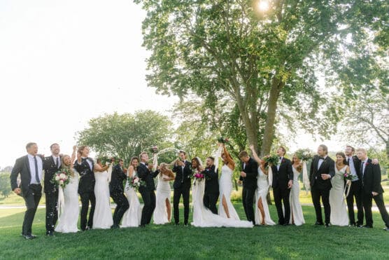 A joyful wedding party, with bridesmaids in white dresses and groomsmen in black suits, celebrate outdoors on green grass, raising bouquets and champagne under tall trees and bright sunlight. by Justin Salem Meyer