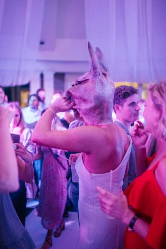 A person in a white dress wears a large pig mask at a lively party, surrounded by other people, some holding drinks and smiling under purple and blue lighting. by Justin Salem Meyer
