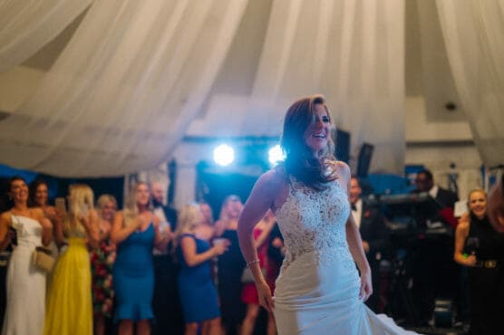 A smiling bride in a white lace wedding dress stands in the foreground, with a group of guests dressed in colorful outfits watching and celebrating behind her under draped white fabric and bright lights. by Justin Salem Meyer