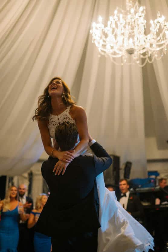 A groom lifts a laughing bride in a white dress during a joyful dance under a chandelier, with guests in formal attire watching and white drapes overhead. by Justin Salem Meyer