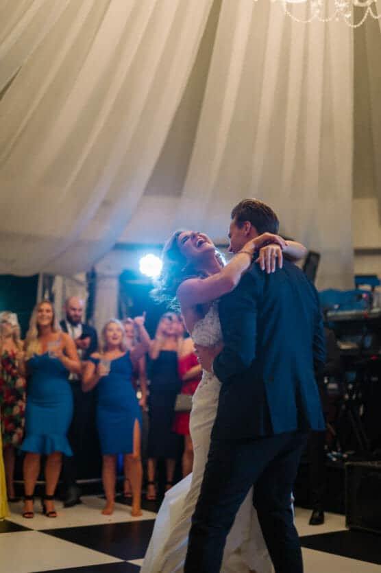 A bride and groom share a joyful first dance under a white tent, surrounded by smiling and clapping guests in formal attire. The bride is laughing with her arms around the groom. by Justin Salem Meyer