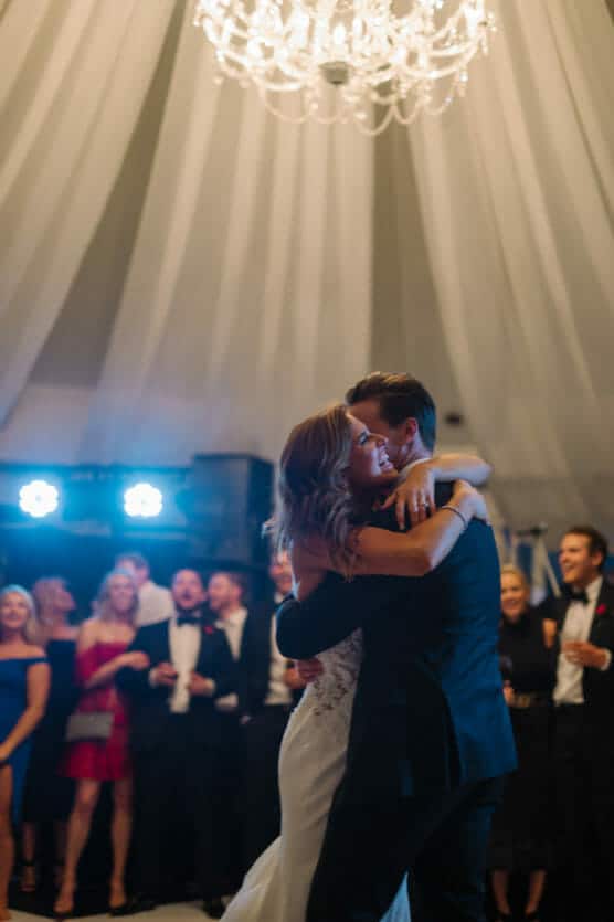A bride and groom share a joyful embrace and dance under a chandelier, surrounded by smiling and applauding guests in formal attire at their wedding reception. by Justin Salem Meyer