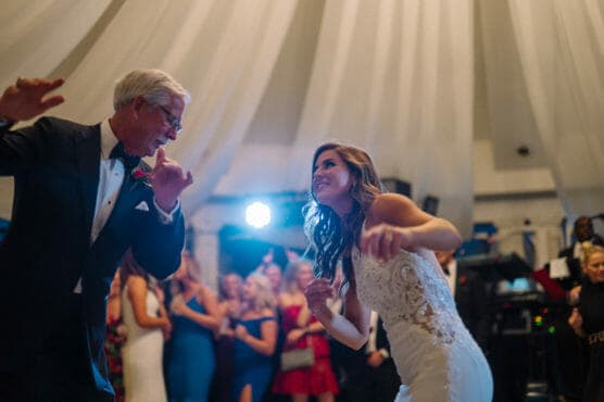 A bride in a white dress and an older man in a tuxedo dance joyfully together under draped white fabric, with a crowd of guests watching and cheering in the background. by Justin Salem Meyer