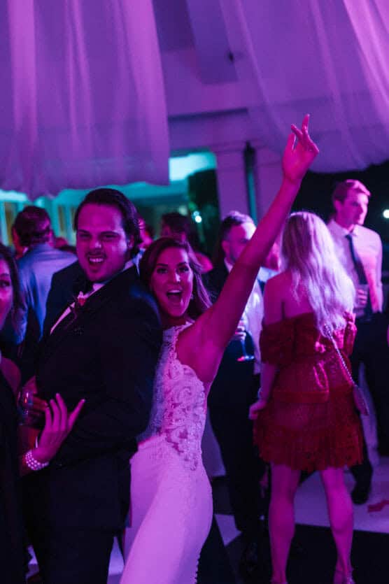 A joyful couple poses playfully at a lively party, with colorful pink and purple lighting. The woman in a white dress raises her arm and smiles, while the man beside her makes a playful face. Other people mingle in the background. by Justin Salem Meyer