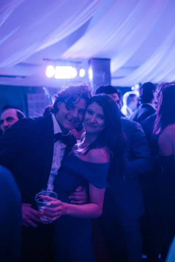 A young couple dressed in formal attire smiles and poses closely together at a lively event under purple-blue lighting, surrounded by other people in party clothes. by Justin Salem Meyer