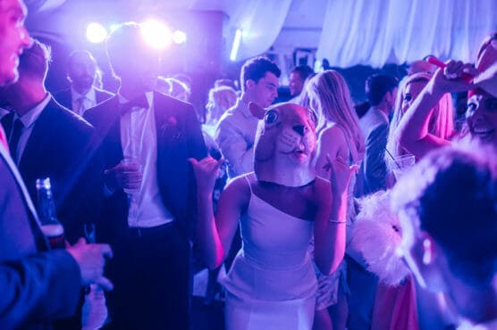 A group of people dressed formally dance at a party with purple lighting. In the center, a person in a white dress wears a squirrel mask and makes a playful hand gesture. The mood is energetic and festive. by Justin Salem Meyer