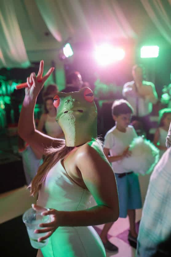 A person in a white dress wears a large, realistic frog mask and makes a peace sign while holding a drink at a party. Colorful lights shine in the background, and several people, including children, are present. by Justin Salem Meyer