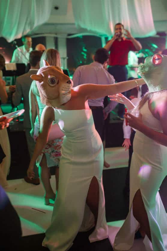 Two people in white dresses and animal masks dance playfully at a lively indoor party, surrounded by other guests. Colorful lights and draped fabric decorate the space, creating a festive atmosphere. by Justin Salem Meyer