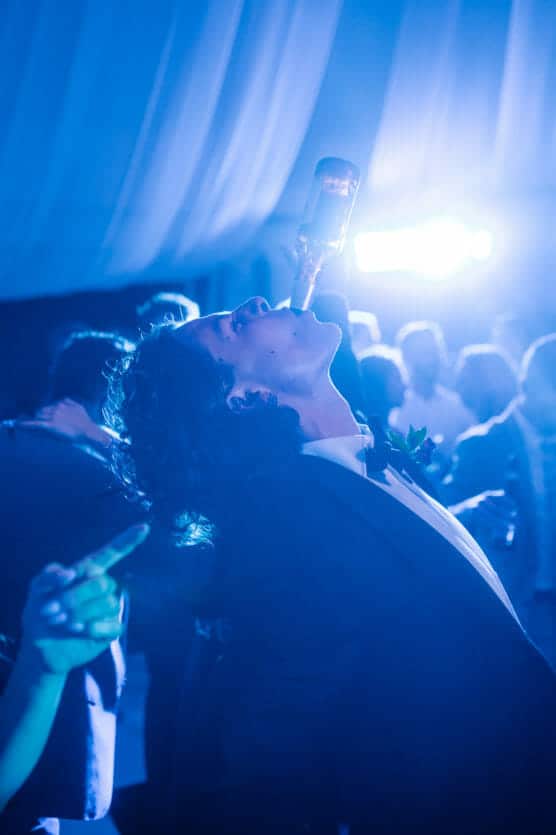 A person in a suit leans back at a lively event, balancing a glass bottle upside down in their mouth, surrounded by people and blue lighting. by Justin Salem Meyer