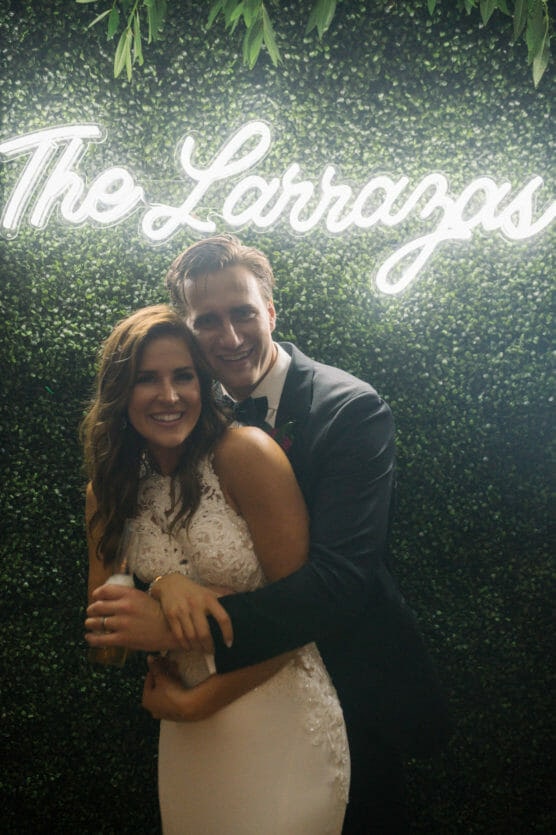 A bride and groom smile and embrace in front of a green ivy wall with a glowing sign that reads “The Larrazas.” The bride wears a white gown and the groom is in a black tuxedo. by Justin Salem Meyer