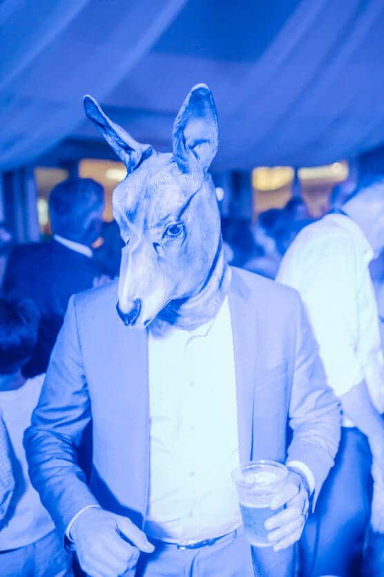 A person wearing a kangaroo mask and a suit holds a drink at a crowded indoor event. The image has a strong blue tint and people in the background are socializing. by Justin Salem Meyer
