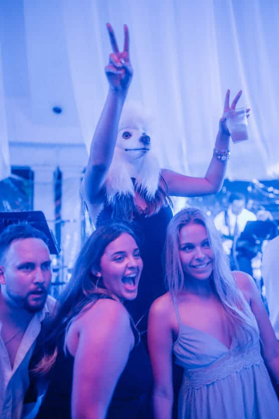 Three people pose and smile at a party while a fourth person behind them, wearing a white animal mask and black dress, raises both arms making peace signs. The scene is lit with blue and purple lights. by Justin Salem Meyer