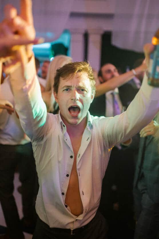 A young man with an open white shirt dances energetically at a party, arms raised and mouth open as if singing or shouting, surrounded by other people enjoying the lively atmosphere. by Justin Salem Meyer
