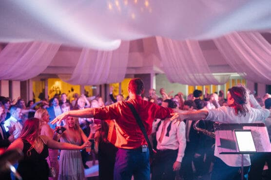 A musician with a guitar and outstretched arms performs on stage facing a lively, dancing crowd at an indoor event with draped white fabric and colorful lighting overhead. by Justin Salem Meyer