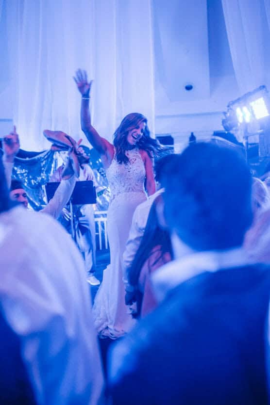 A bride in a white gown smiles and raises her arm while being lifted on a chair during a lively wedding celebration, surrounded by guests under blue lighting. by Justin Salem Meyer