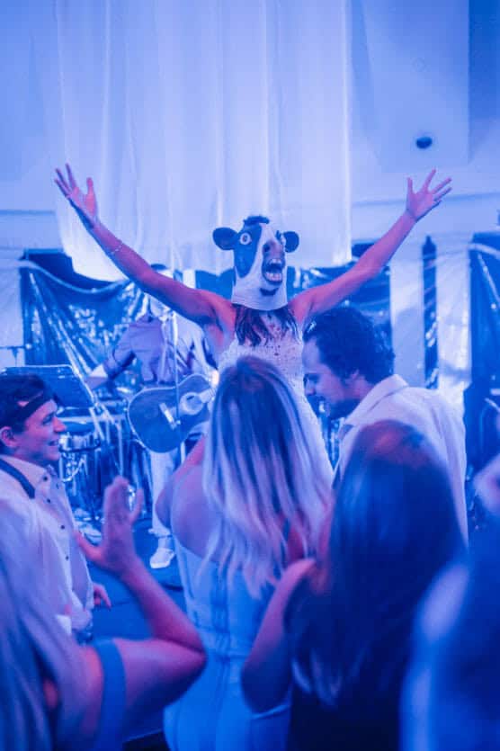 A person wearing a cow mask and a white dress stands with arms raised on a stage, surrounded by people dancing at a lively party bathed in blue lighting. by Justin Salem Meyer