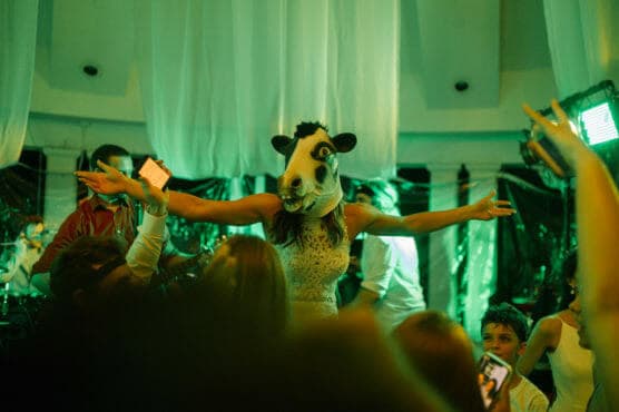 A person wearing a cow mask and a white dress stands with arms outstretched at a lively party with green lighting, surrounded by people taking photos and celebrating. by Justin Salem Meyer