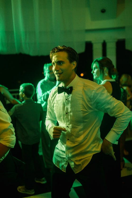 A man in a white dress shirt and black bow tie dances energetically at an indoor event, surrounded by other guests under green lighting. by Justin Salem Meyer