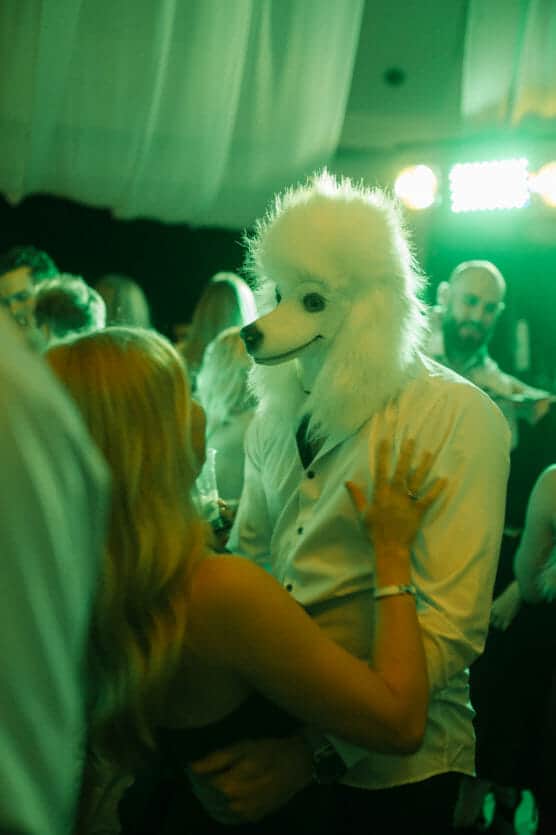 A person wearing a fluffy white poodle mask and a white shirt dances closely with another person at a party, surrounded by other guests under green lighting. by Justin Salem Meyer