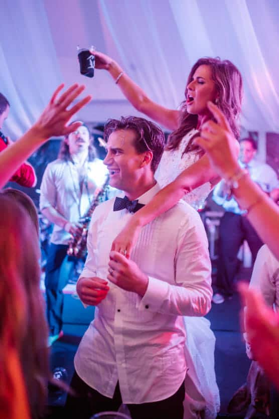 A bride sits on a grooms shoulders, raising a drink and cheering at a lively wedding reception with guests dancing and a band performing in the background. by Justin Salem Meyer