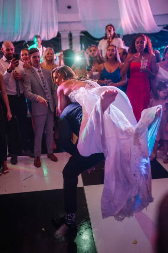 A bride in a white gown is being lifted playfully by a man on a dance floor, surrounded by cheering and smiling wedding guests. The scene is festive with colorful lights and draped fabric on the ceiling. by Justin Salem Meyer