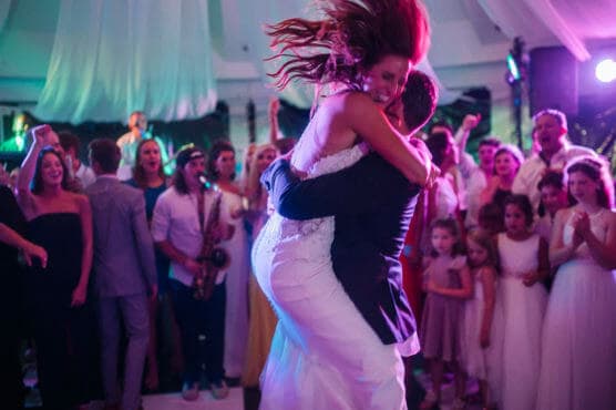A bride joyfully leaps into a grooms arms as they embrace on the dance floor, surrounded by cheering and smiling wedding guests under colorful lights. by Justin Salem Meyer