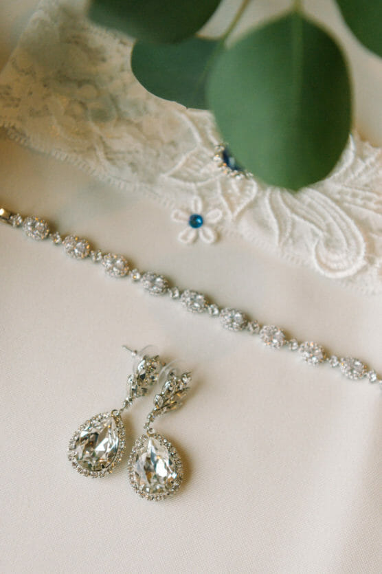 A pair of teardrop-shaped crystal earrings, a sparkling bracelet, and a piece of white lace with a small blue flower detail are arranged on a light-colored surface with green leaves partially visible above. by Justin Salem Meyer