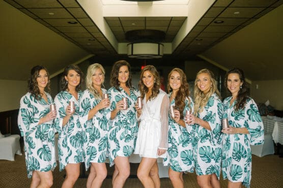 Eight women, one in a white robe and seven in matching green leaf-patterned robes, stand together indoors, smiling and holding drinks, as if celebrating before a special event. by Justin Salem Meyer