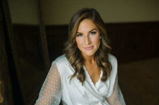 A woman with long, wavy brown hair wearing a white robe with sheer, dotted sleeves looks at the camera and smiles softly, seated indoors against a neutral background. by Justin Salem Meyer