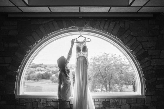 A woman reaches up to touch a wedding dress hanging in front of a large arched window, with a scenic view of trees and fields outside. The image is in black and white. by Justin Salem Meyer