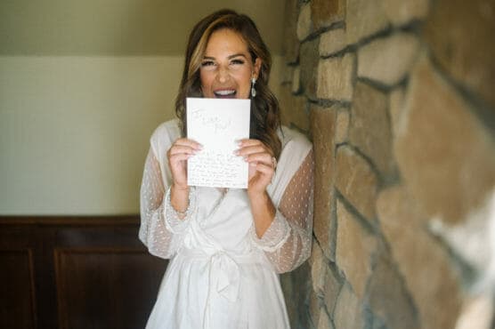 A woman in a white dress smiles joyfully while holding up a handwritten letter that says I love you! in front of her face, standing indoors next to a stone wall. by Justin Salem Meyer